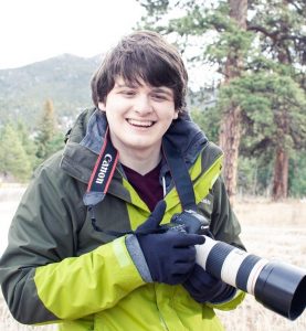 Ein Mann in einer grünen Jacke hält eine Kamera und lächelt in der Natur